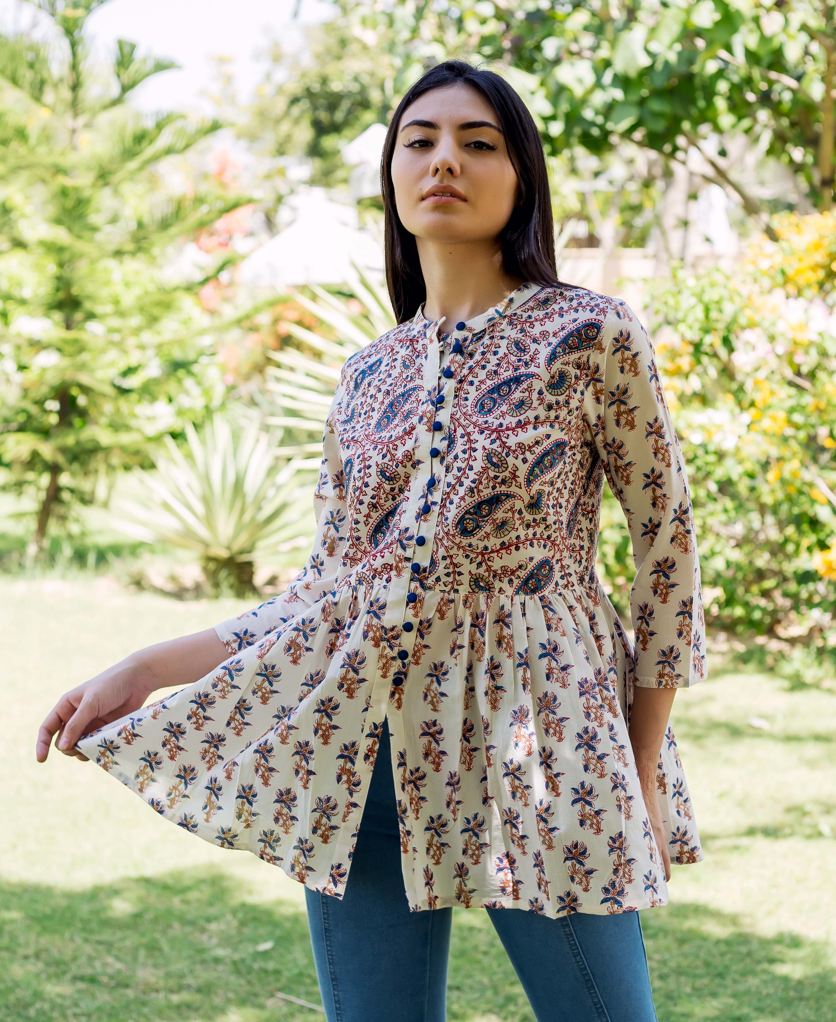 Model wearing a cream peplum top with blue, red, and brown ethnic patterns in a garden.