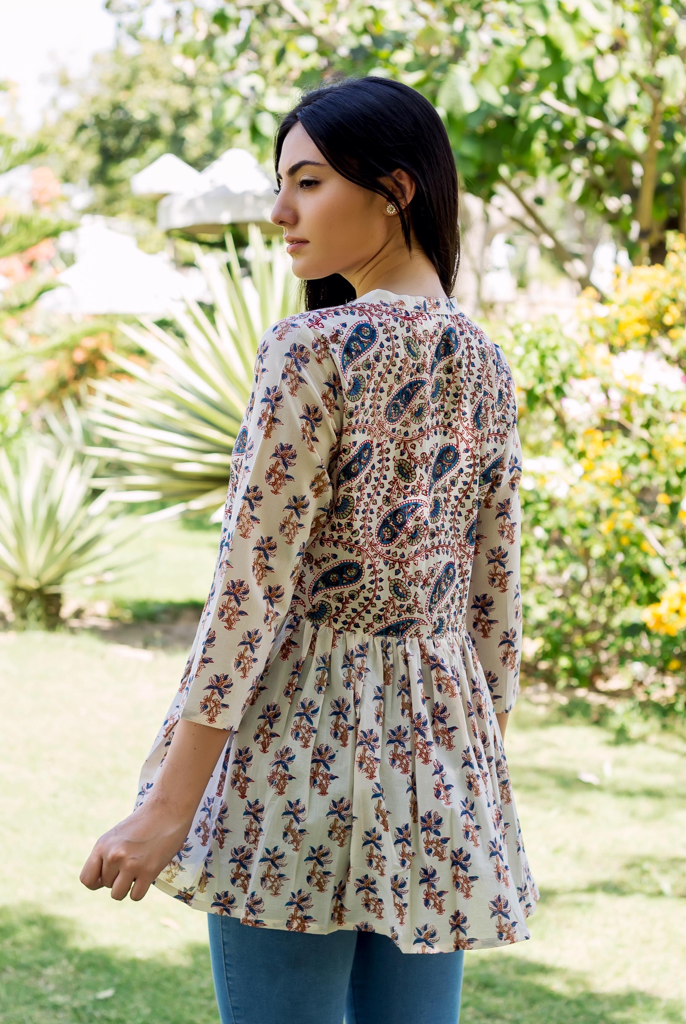 Model wearing white peplum top with blue, brown paisley and floral prints, facing away.