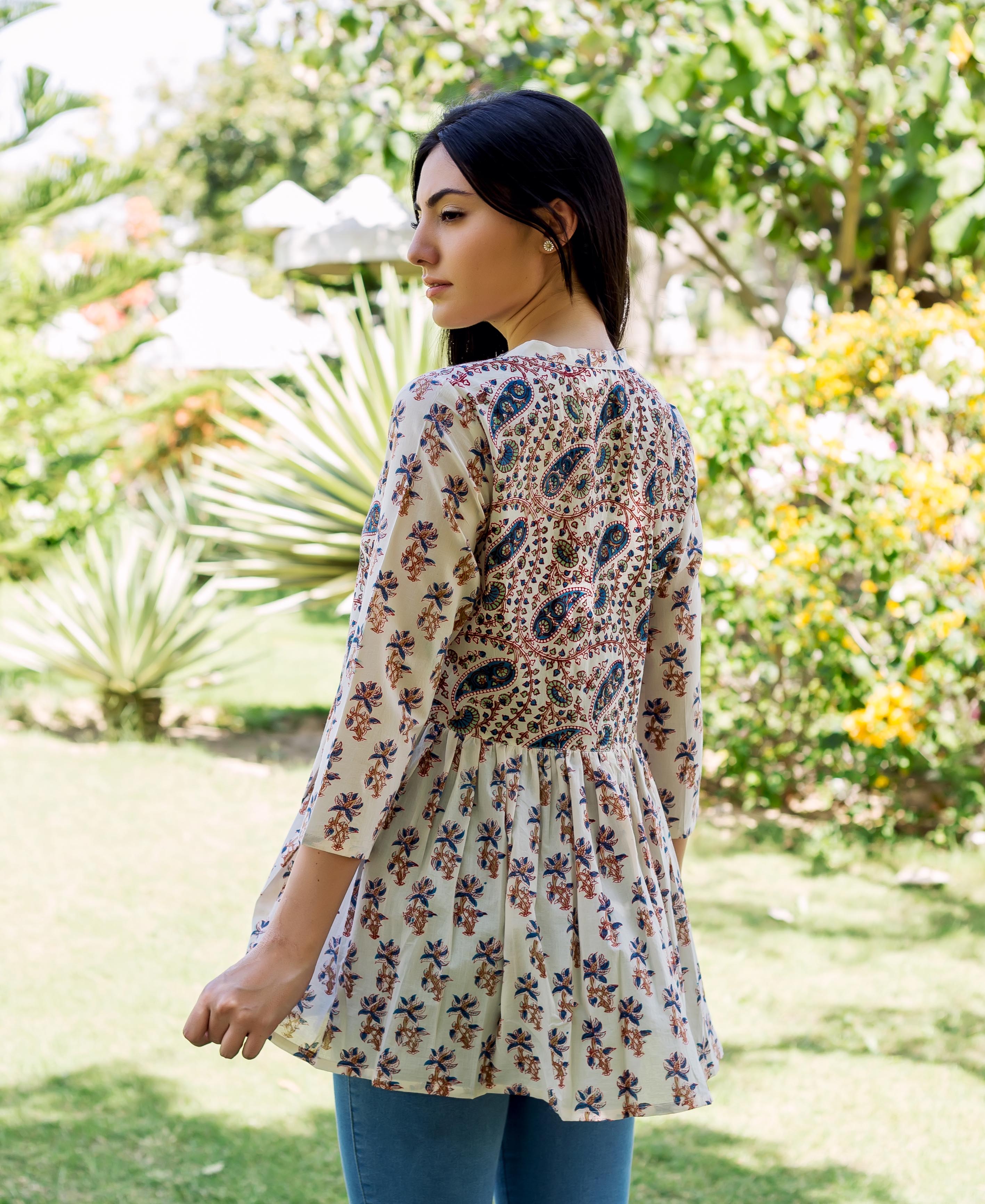Model wearing white peplum top with blue, brown paisley and floral prints, facing away.