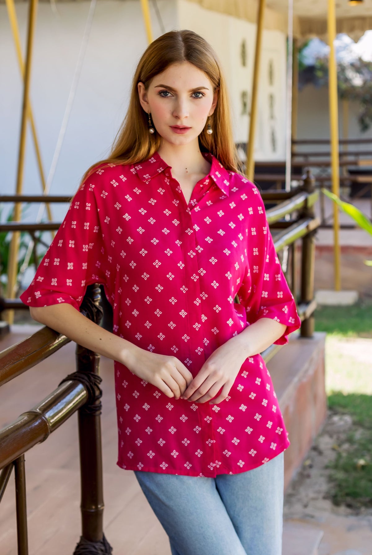 Young woman models a magenta button-down shirt with white bandhej print, leaning on railing.