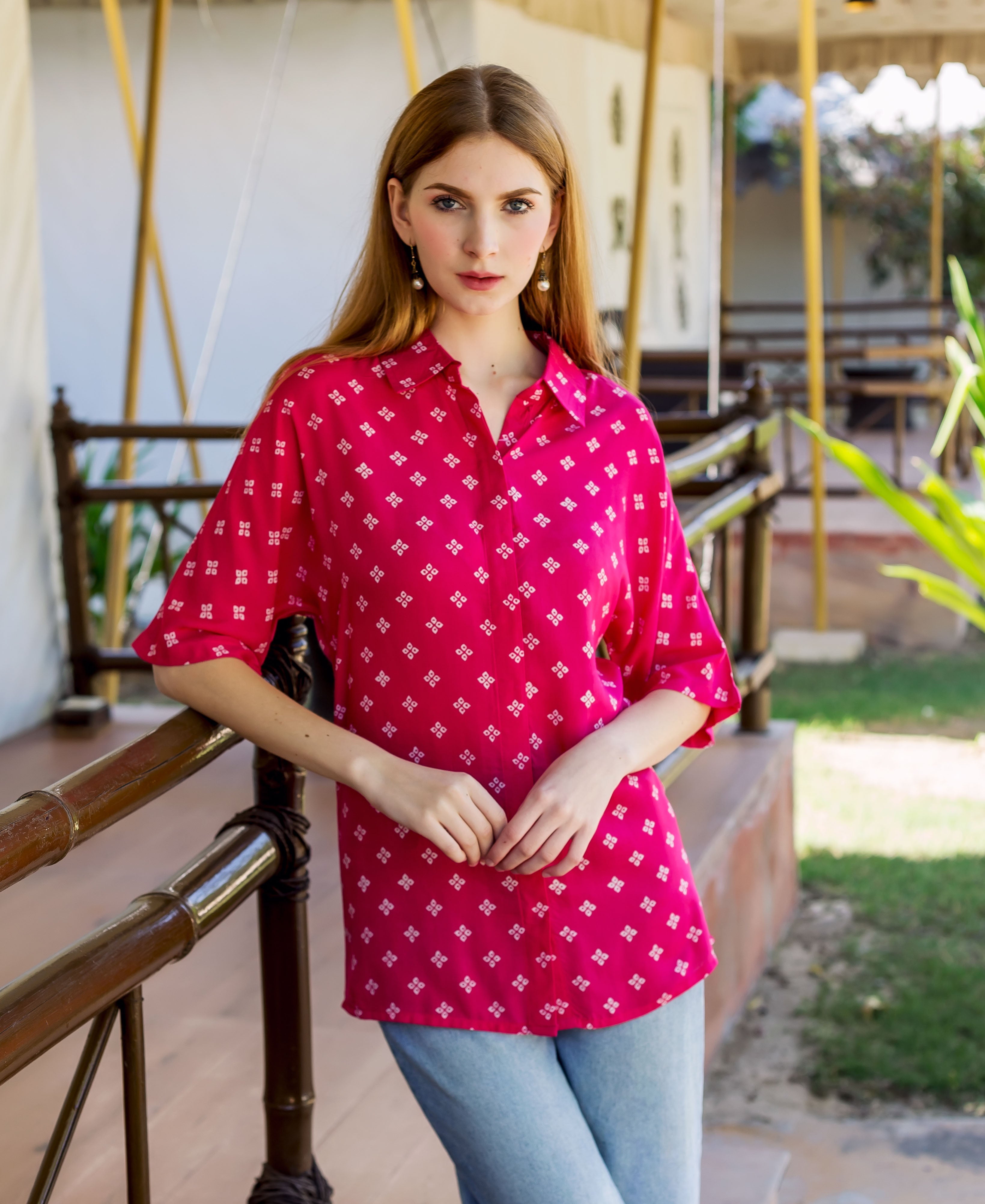 Young woman models a magenta button-down shirt with white bandhej print, leaning on railing.