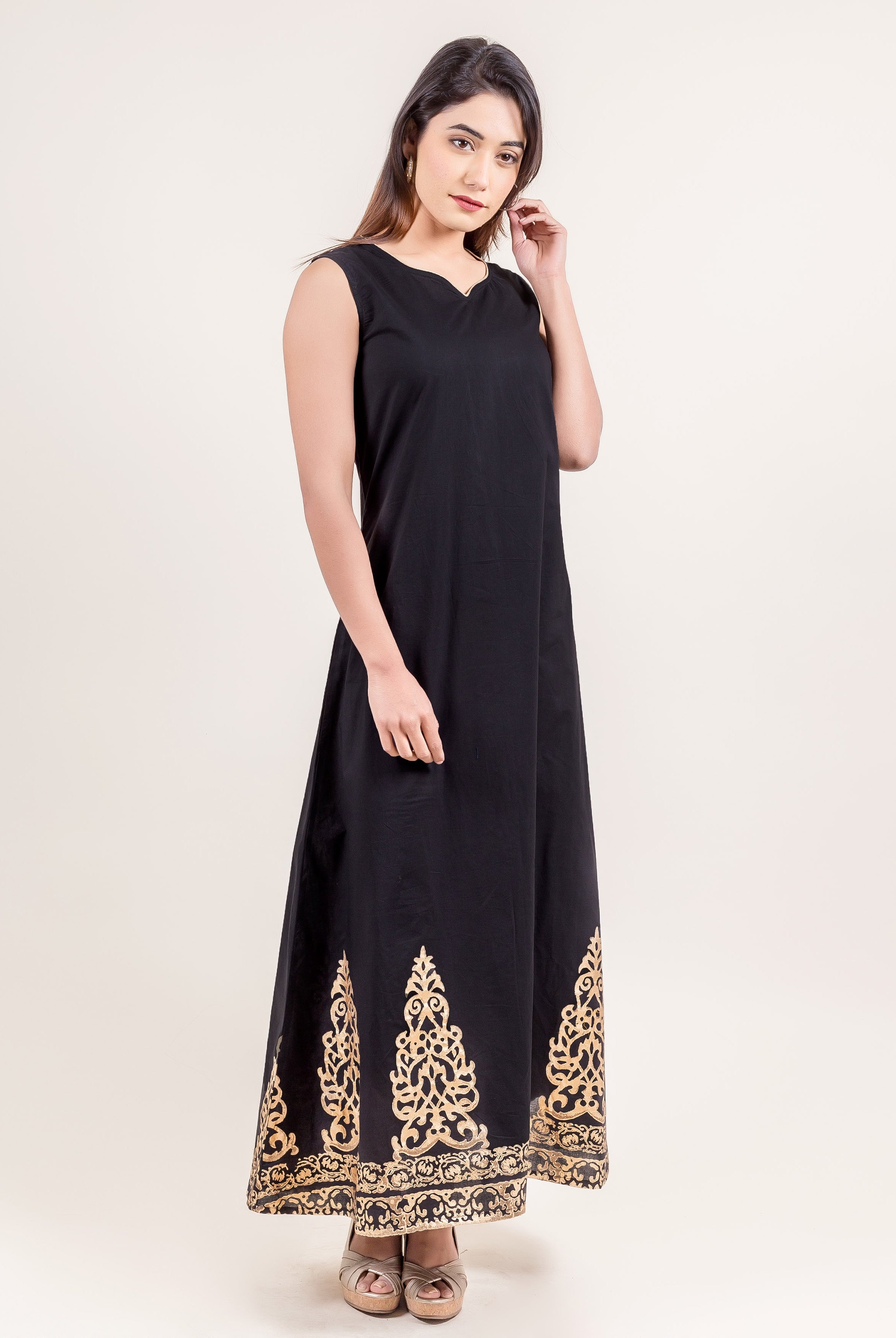 Model in a black sleeveless long dress with ornate gold ethnic print at the hem.