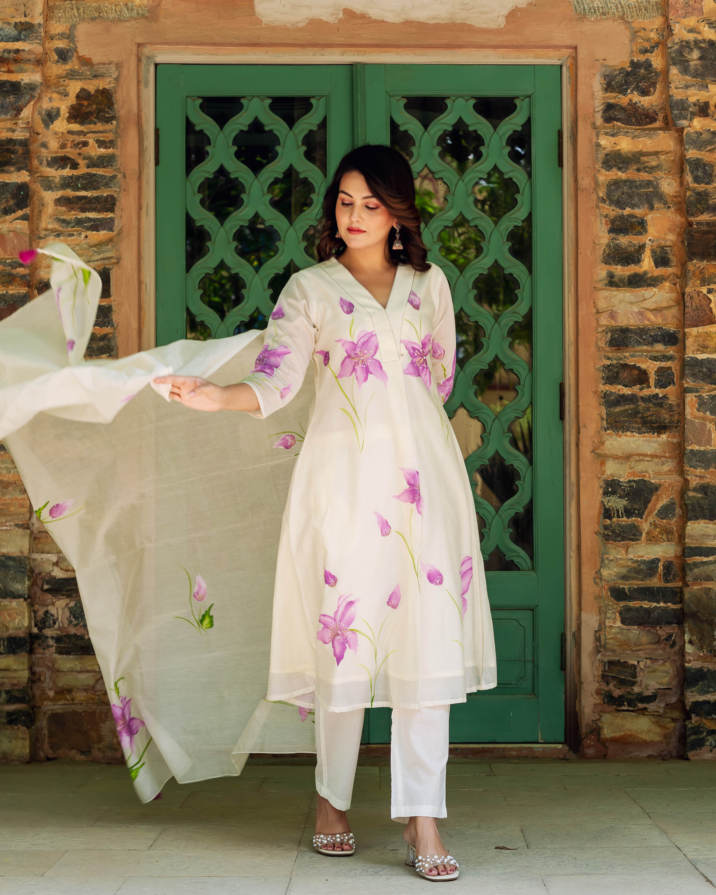 A woman in a white kurta with purple flowers and green stems, standing in front of a green door.