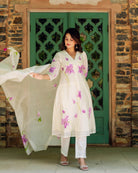 A woman in a white kurta with purple flowers and green stems, standing in front of a green door.