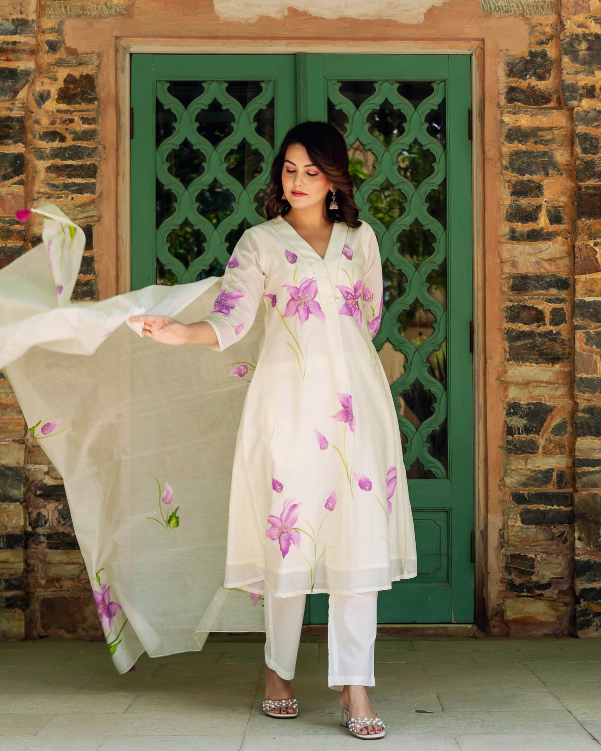 A woman in a white kurta with purple flowers and green stems, standing in front of a green door.