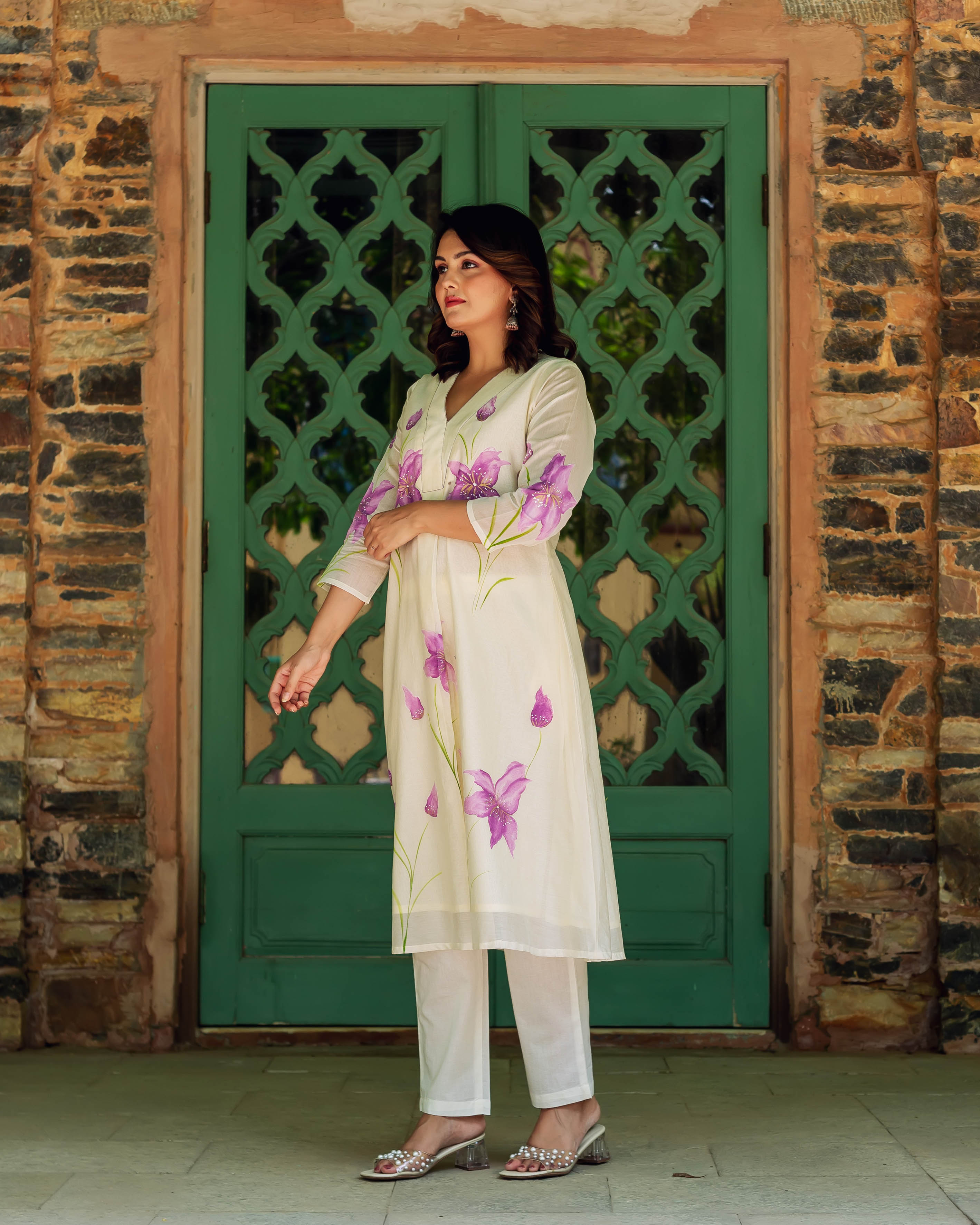 A woman in an ivory kurta with purple flowers, standing in front of a green door.