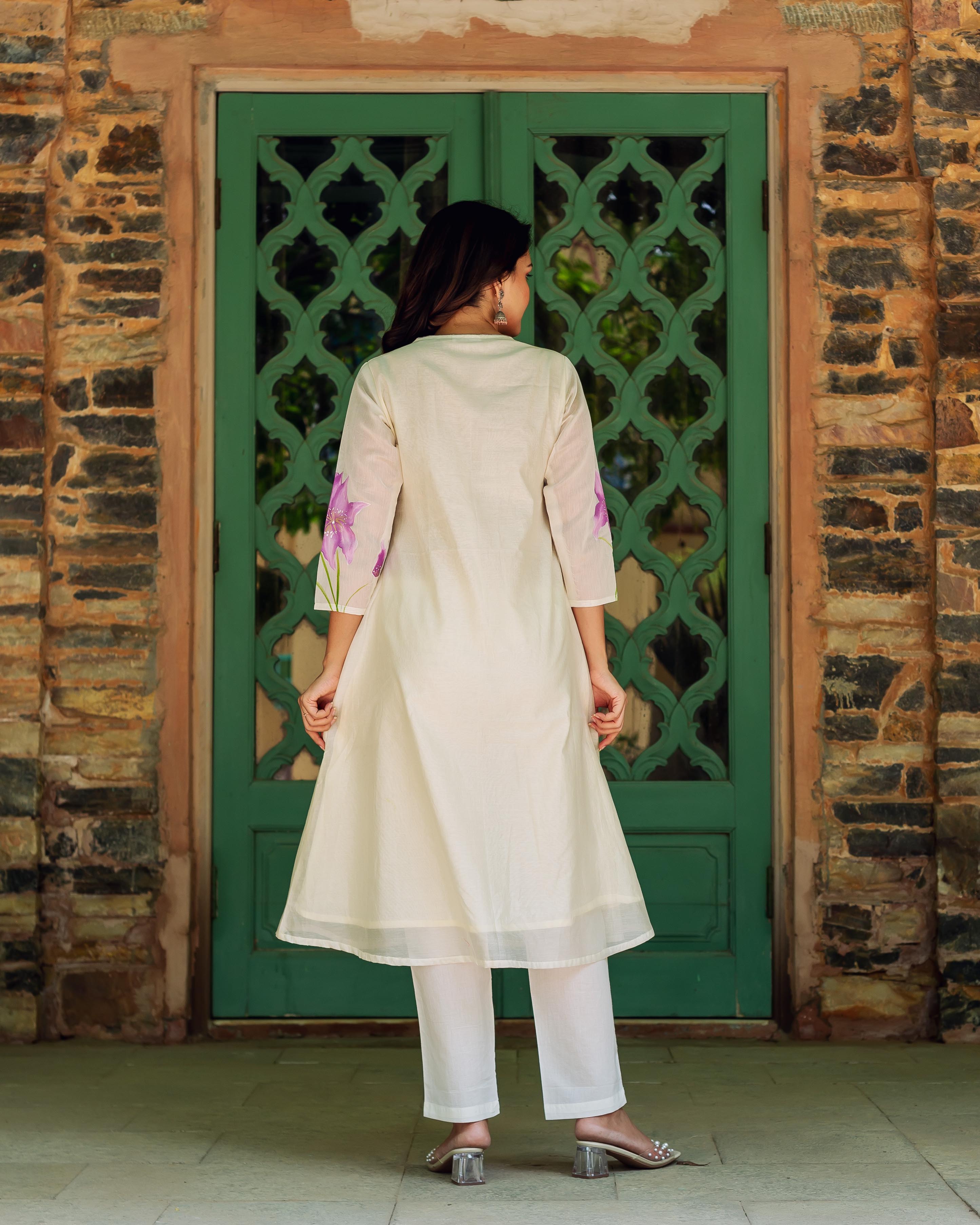 Woman in cream kurta with purple blossoms on sleeves, standing before green door.