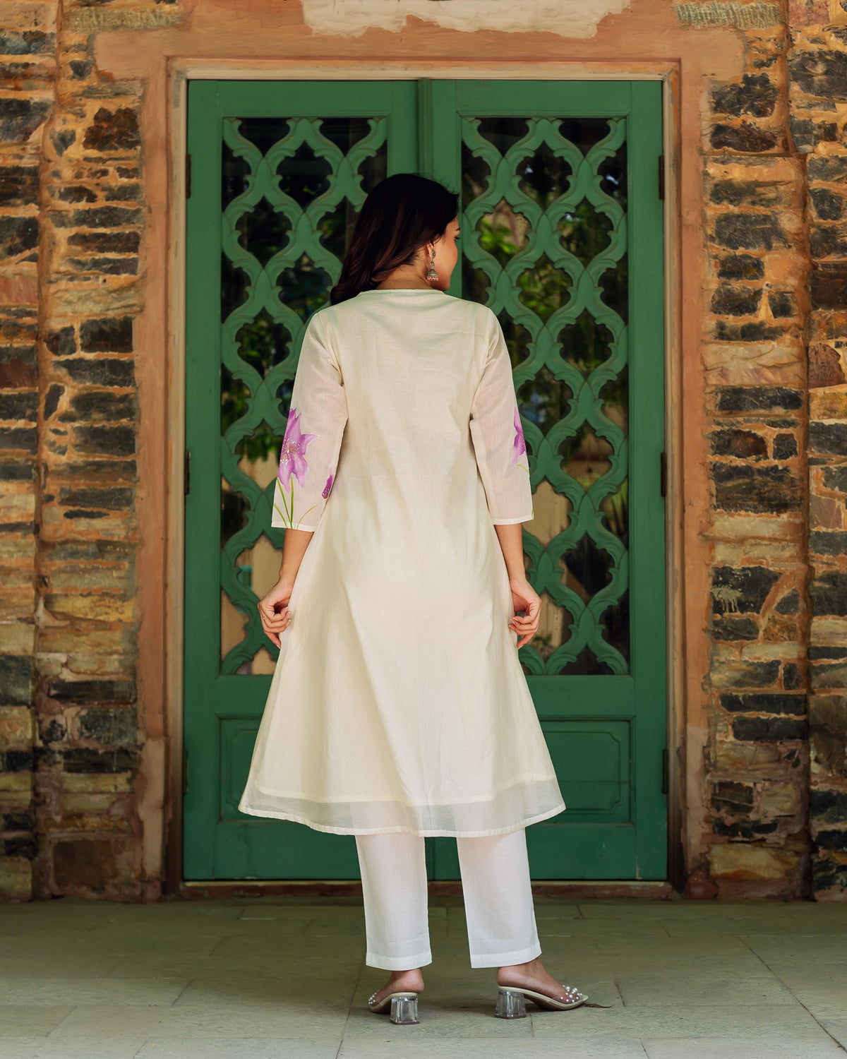 Woman in cream kurta with purple blossoms on sleeves, standing before green door.