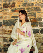 Woman in ivory kurta with purple floral print, sitting against stone wall.