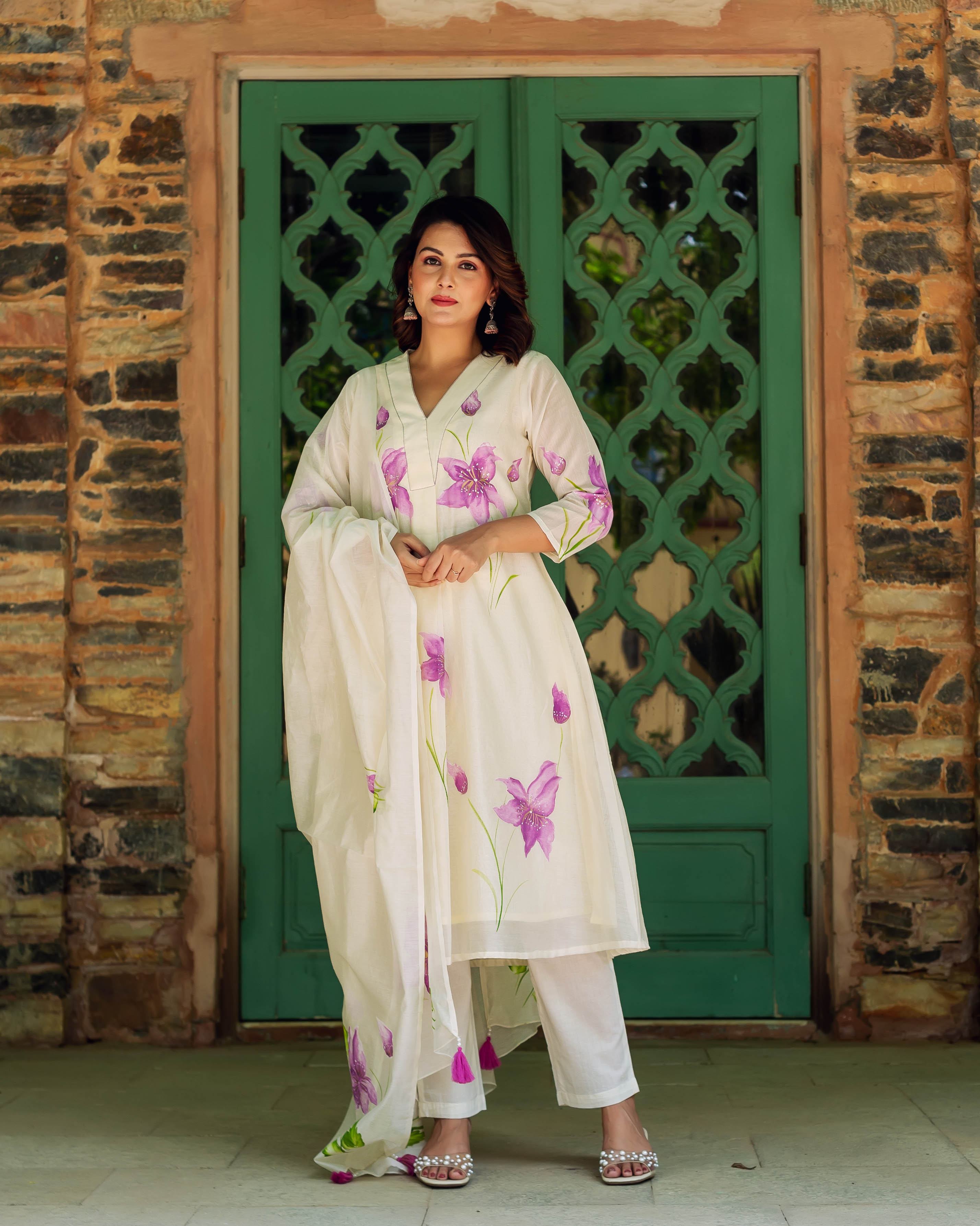 A woman in a white kurta with purple flowers and green stems, standing in front of a green door.
