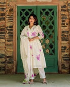 A woman in a white kurta with purple flowers and green stems, standing in front of a green door.